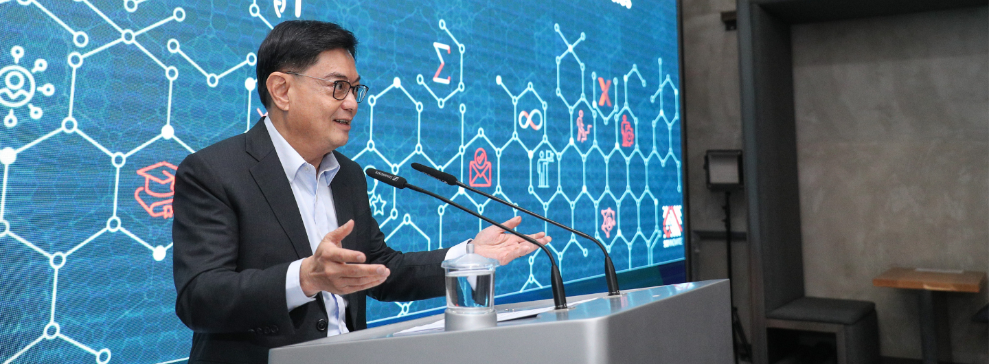 Man at podium gestures during presentation; blue screen with icons and network diagram background.
